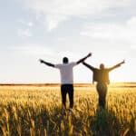 Two people walking through cornfield at sunset with hands up.