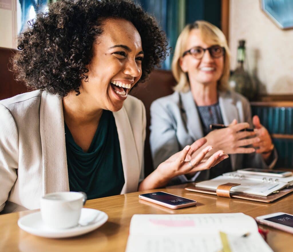 Two women smiling and laughing at table with planners and cellphones in front of them.