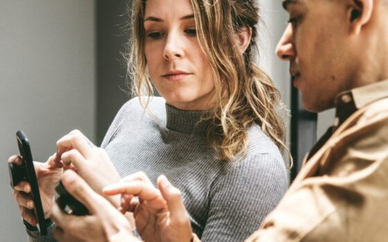 Woman and man sitting next to one another, looking and conversing about their cell phones.