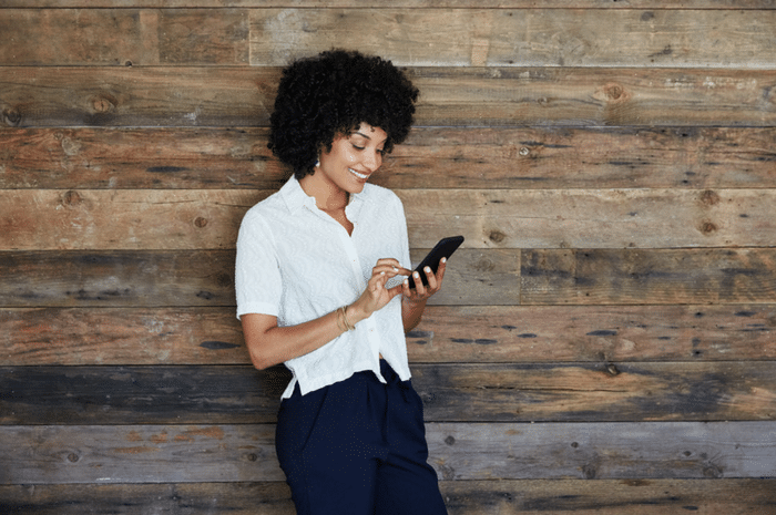 African American woman smiling standing against wood wall using her phone.